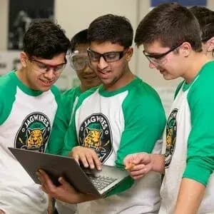 Group of students wearing matching robotics club raglan t-shirts and safety glasses look at a laptop.