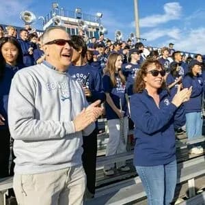 Parents wearing custom school crewneck sweatshirt and quarter zip sweatshirt stand in a crowded stand as a sporting event.