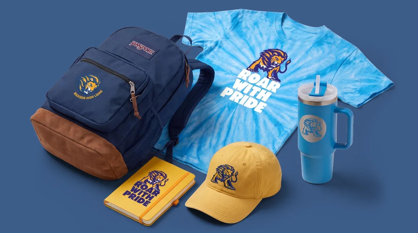 Custom school-branded merchandise displayed together, including a backpack, tie-dye T-shirt, notebook, baseball cap, and insulated tumbler featuring a matching lion mascot logo and “Roar With Pride” design.