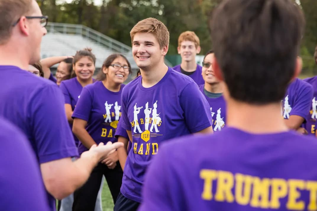 Students wearing matching custom purple marching band t-shirts gather and laugh during practice.