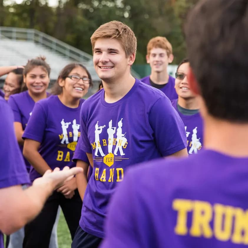 Students wearing matching custom purple marching band t-shirts gather and laugh during practice.