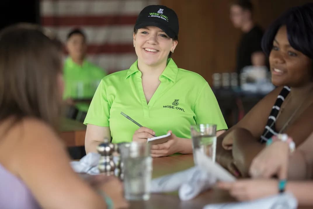 A server wearing branded hat and polo shirt, takes a customer order in the Wise Owl American Bistro.