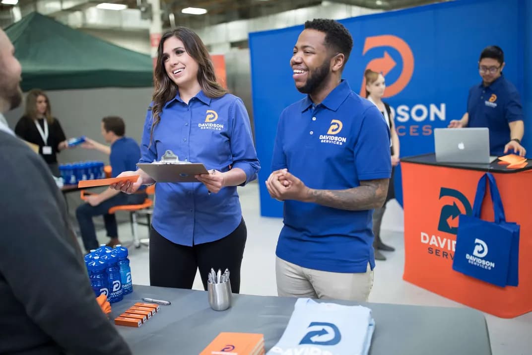 Two coworkers wearing coordinating branded uniforms, talk to a prospective client at their booth featuring promotional giveaways and custom signage.