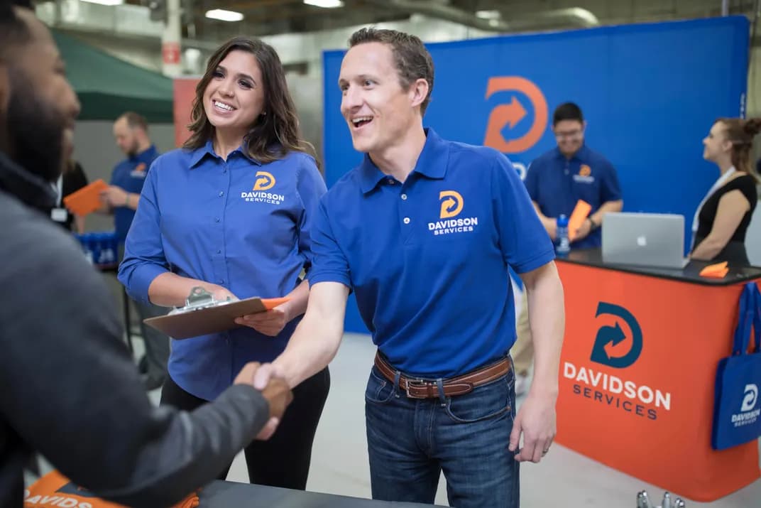 Two coworkers wearing coordinating brand uniforms talk to a prospective client at their tradeshow booth.