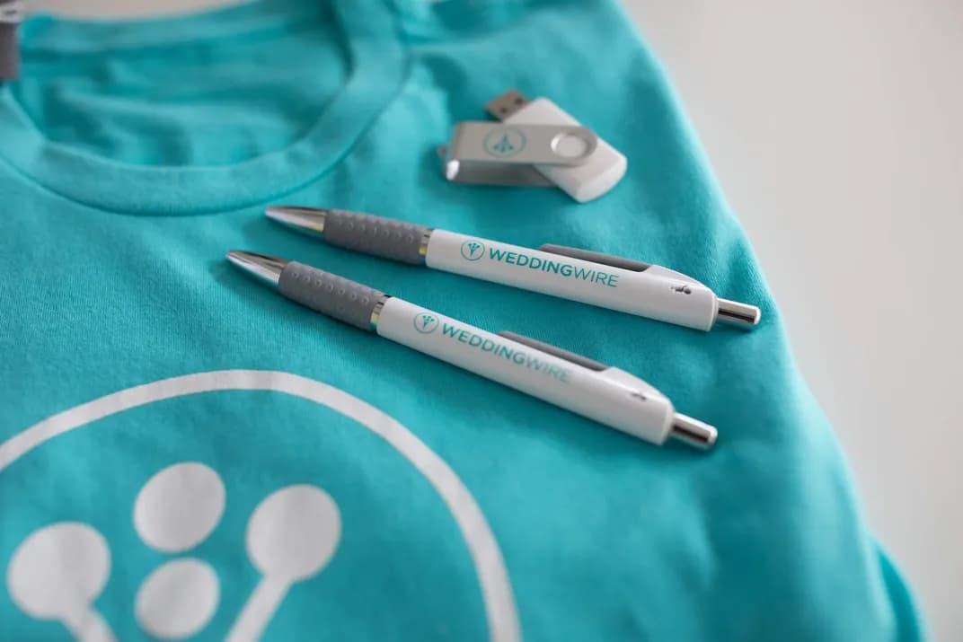 Custom branded WeddingWire pens, USB drive, and teal t-shirt with company logo.