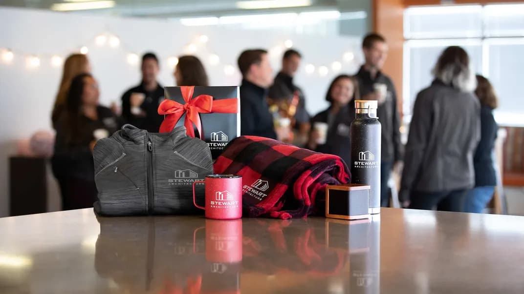 Branded company gifts including custom logo jacket, mug, blanket, and bottle displayed on a table with coworkers in the background.