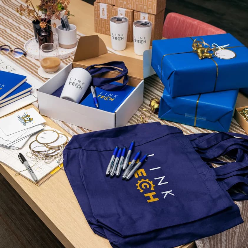 Desk with custom branded Link Tech notebooks, mugs, pens, tote bags, and wrapped gift boxes prepared for corporate gifting.