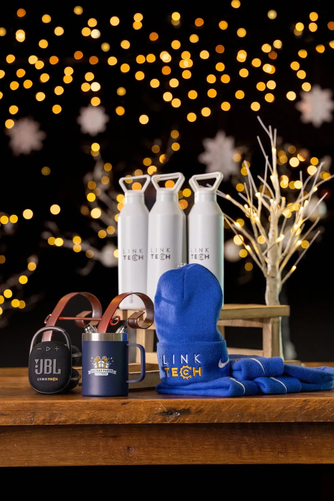 Branded company holiday gifts including custom logo water bottles, mug, beanie, and speaker displayed with festive lights