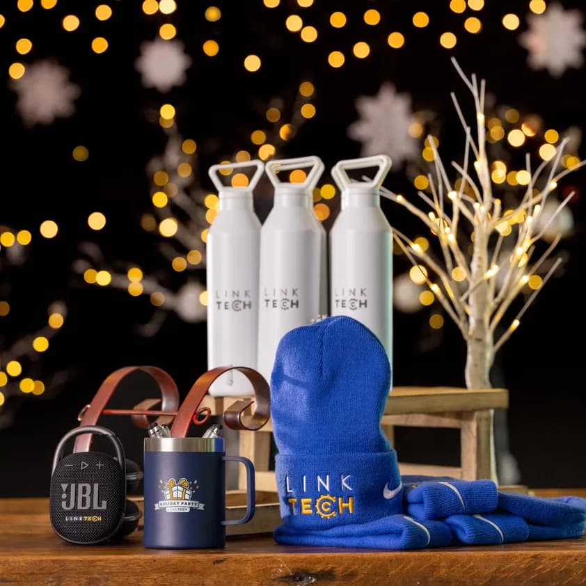 Branded company holiday gifts including custom logo water bottles, mug, beanie, and speaker displayed with festive lights