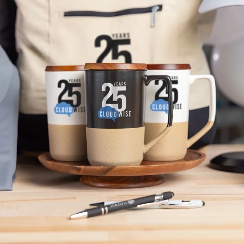 Custom branded mugs and pens featuring the Cloudwise 25 years anniversary logo displayed on a wooden tray