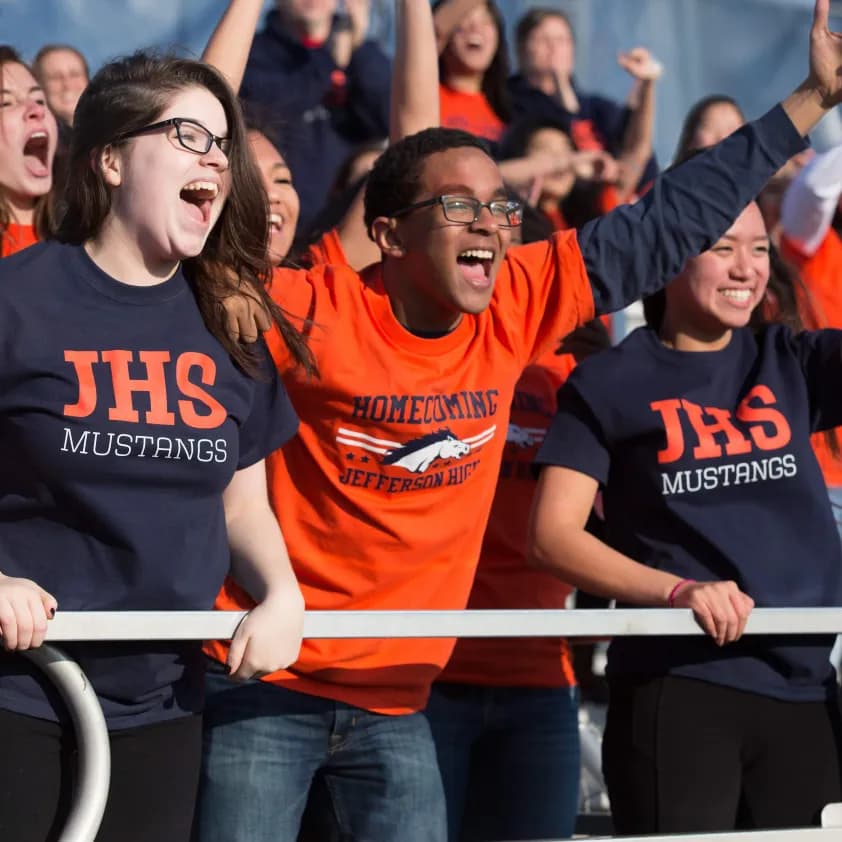 High School students cheer in a crowded stand wearing custom spirit wear t-shirts.