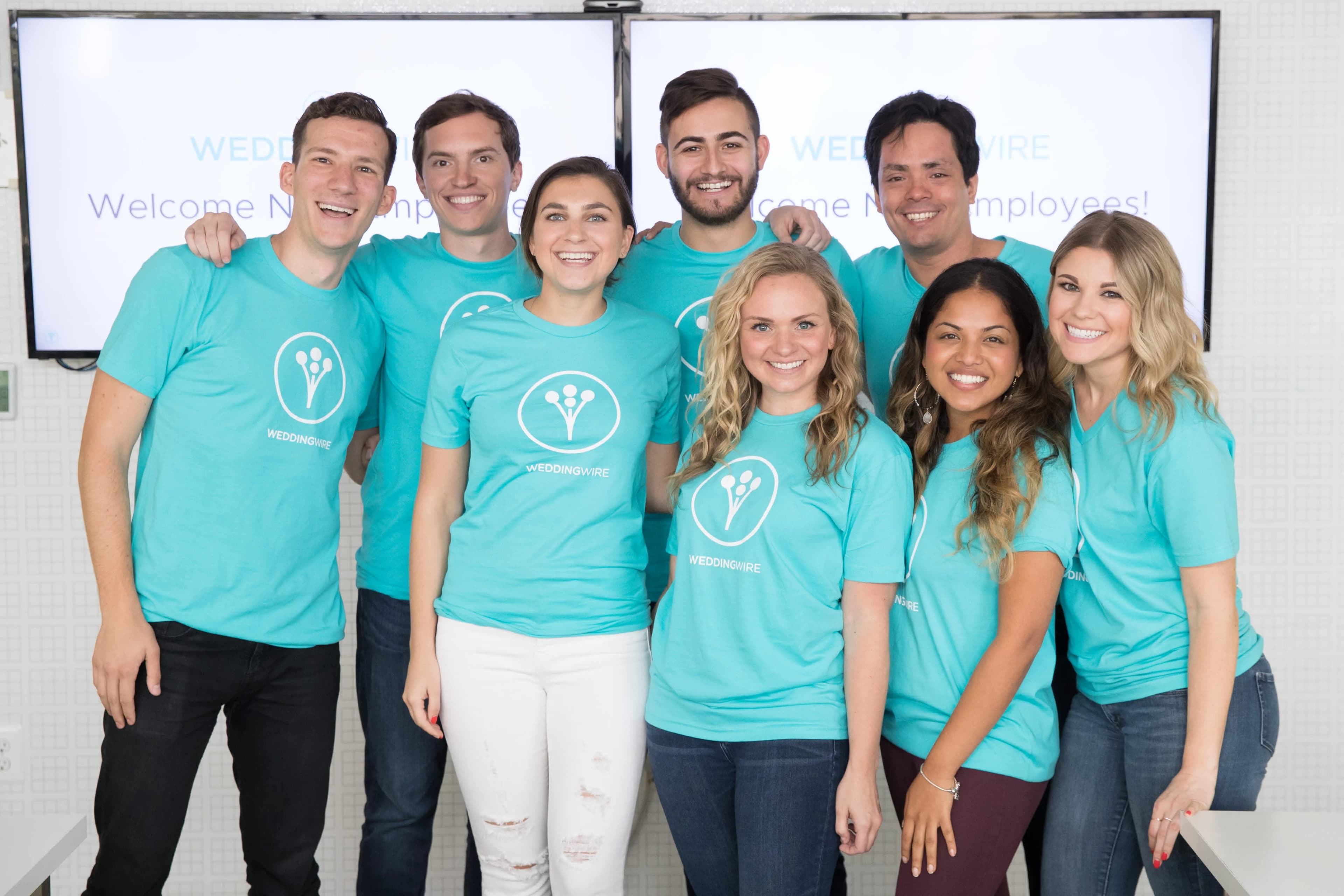 Group of employees wearing matching custom teal WeddingWire t-shirts smiling together in an office setting