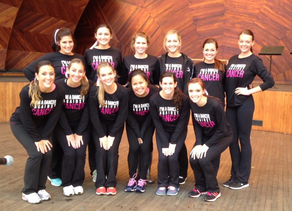 A group of dancers poses on stage wearing matching custom t-shirts that say, "I'm a dancer against cancer."