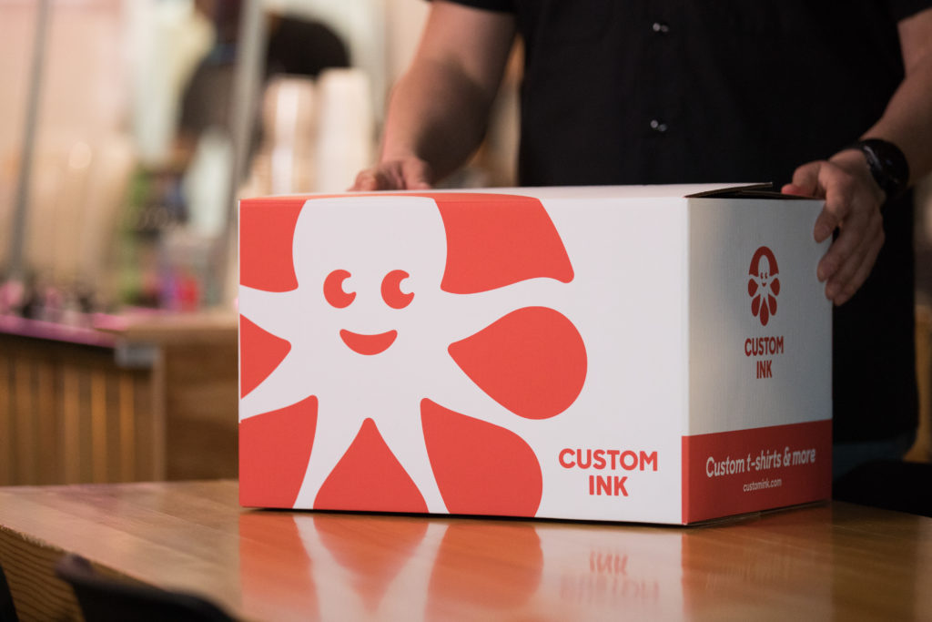 Custom Ink box on a table in foreground with figure holding it in the background.