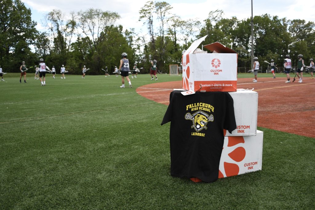 A t-shirt hangs from a pile of Custom Ink boxes while students play lacrosse in the background.
