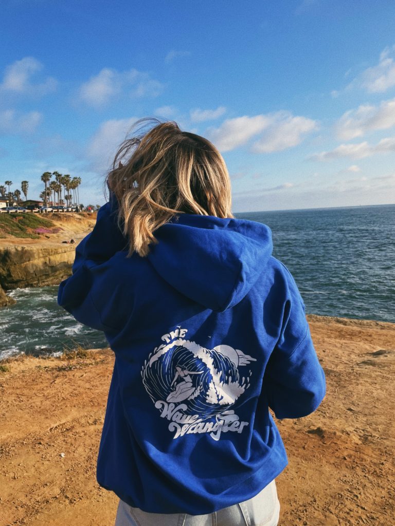 Woman standing in front of the beach wearing a hoodie with The Wave Wrangler printed on the back