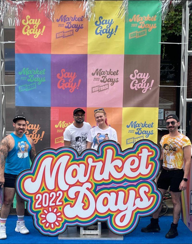 Skylar and others stand behind a sign reading "2022 Market Days" and a backdrop of squares decorated with the phrase "Say Gay!"