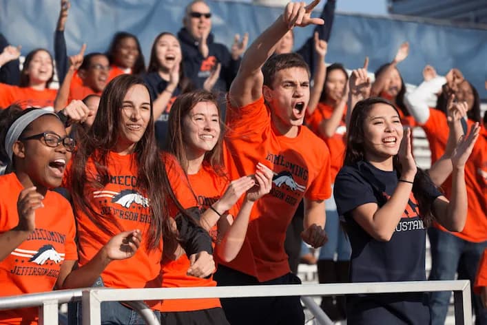 High school students wearing matching Homecoming Jefferson High orange t-shirts cheering in a grandstand.
