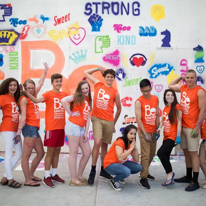 Group of students wearing matching custom orange t-shirts posing in front of a colorful mural.