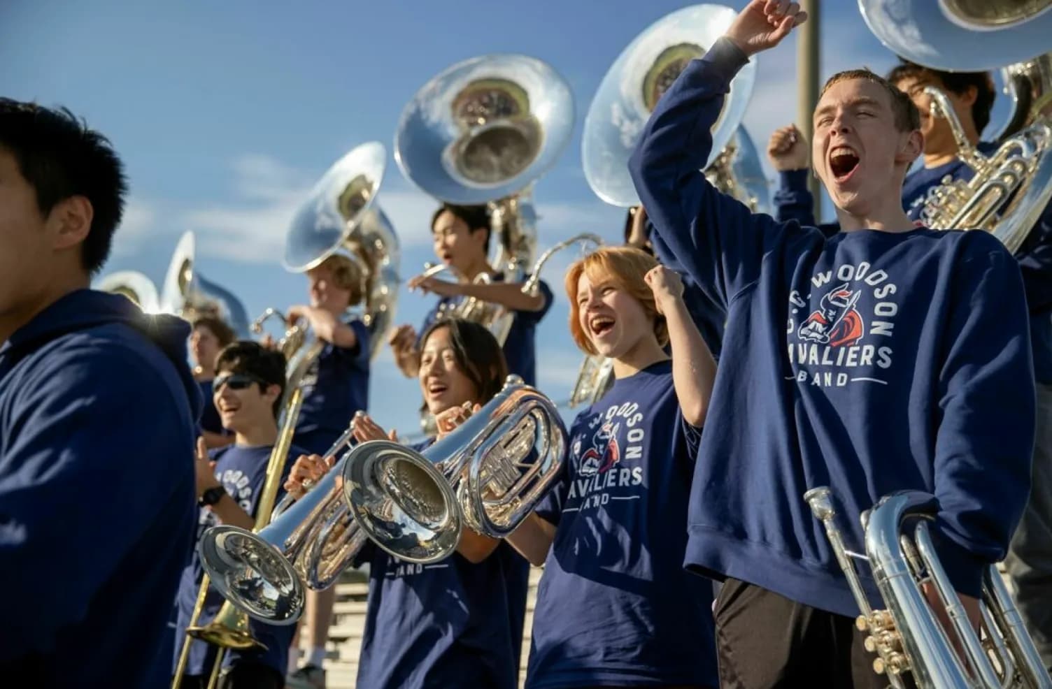 High school marching band wearing matching custom logo sweatshirts cheering and playing instruments in the stands.