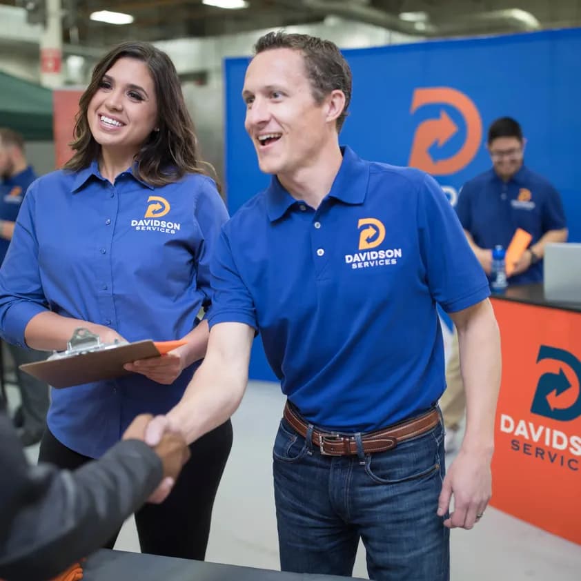 Two coworkers wearing coordinating brand uniforms talk to a prospective client at their tradeshow booth.