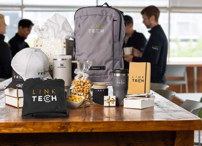 Coworkers wearing custom t-shirts and zip-ups gather around a table containing customized t-shirts, corporate gifts, including Stanley drinkware, a backpack, a hat, a scarf, a notebook with a pen, a Bluetooth speaker, and snacks.